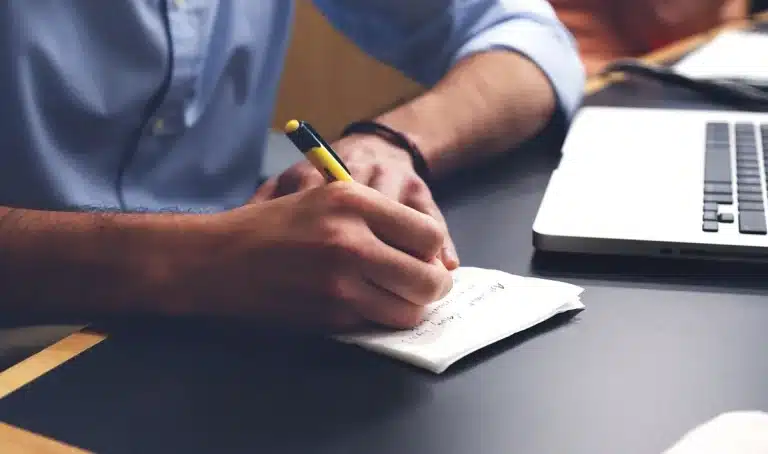 Daylit home office desk with laptop showing an email app, hand typing and a notepad